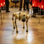 Robotyczny pies idzie między czerwonymi krzesłami w nowoczesnym wnętrzu